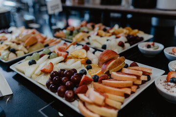 A beautifully arranged assortment of fresh, sliced fruits including apples, oranges, grapes, strawberries, and melons displayed on white serving platters at a buffet, creating a vibrant and healthy 