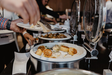 A buffet-style catering service with guests serving themselves from chafing dishes filled with gourmet food, including a creamy dish garnished with grilled lemon slices, in a professional event 