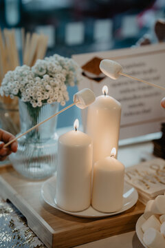 A close-up of a cozy wedding dessert station featuring marshmallows being roasted over elegant white pillar candles, complemented by delicate floral decor