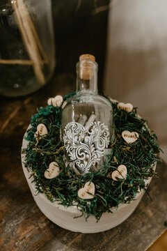 A beautifully decorated glass bottle with a white intricate heart design, sealed with a cork, placed on a bed of green moss in a wooden bowl adorned with small wooden heart-shaped tags featuring 