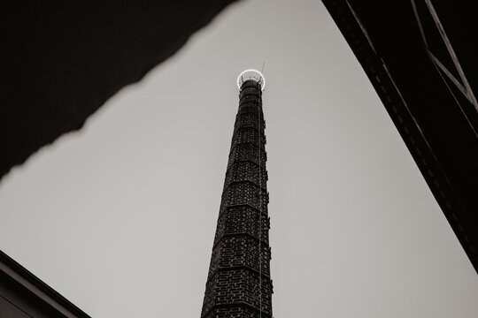 A towering industrial brick chimney with a metal ladder running along its side, photographed from a low-angle perspective between two dark structures, creating a dramatic and symmetrical composition 