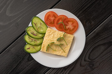 A white plate with an omelette, sliced cucumber, and tomatoes garnished with microgreens