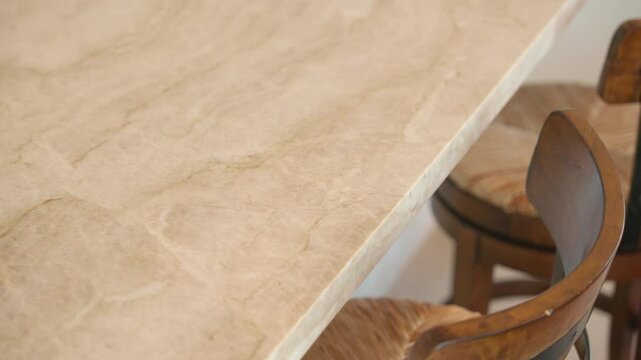 Close up view of light colored marble or stone tabletop with smooth surface. Below tabletop there are wooden chairs with curved backs and cushioned seats
