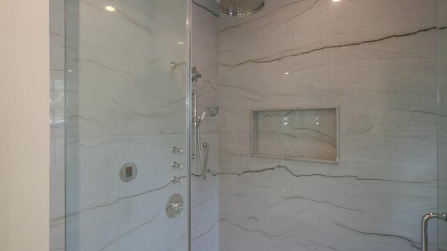 Modern shower area with glass doors and marble walls. Shower has built in niche for storage, rainfall showerhead and handheld shower head