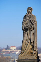 Fototapeta premium The statue shows a woman wearing a hooded robe, with a calm and pensive expression, set against a bright blue sky and distant buildings