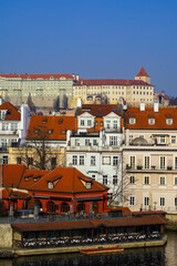 The view shows colorful rooftops of Prague with the castle complex in the background under a bright blue sky