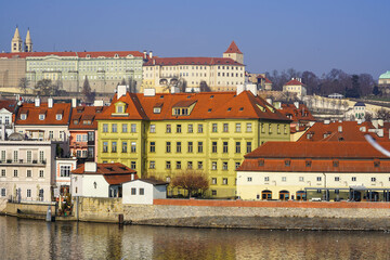 Fototapeta premium A peaceful scene of colorful buildings with red rooftops along a riverbank, and in the distance, a castle complex on a hill