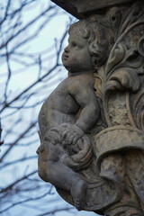 A finely sculpted stone cherub with delicate features and curly hair is carved into an ornate architectural structure, standing against a backdrop of bare tree branches