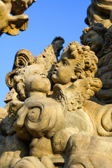 A beautifully crafted stone sculpture features cherubs with expressive faces and delicate wings, bathed in warm sunlight against a clear blue sky