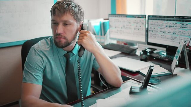 The tired dispatcher is talking on the phone. A busy office worker jugles several tasks on the computer table while working on the phone.
