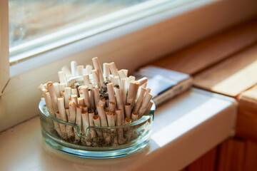 An overflowing ashtray with cigarettes against a window background symbolizes the fight against addiction and the harmful consequences of smoking.