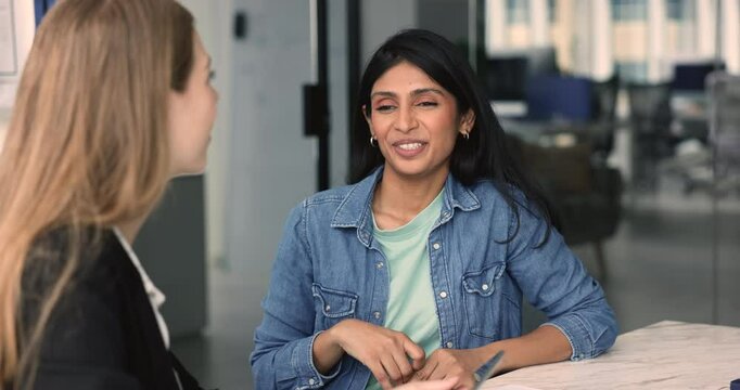 Positive young Indian project manager talking to female colleague at office table, listening to speaking co-worker, enjoying professional networking, corporate communication, discussing business tasks