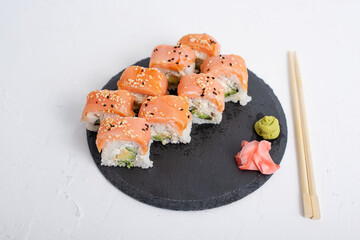 Plate of fresh salmon sushi Philadelphia rolls garnished with sesame seeds, served with wasabi and pickled ginger on a black slate plate. Chopsticks rest beside the dish on a white background.