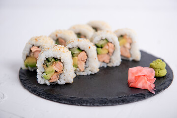Close-up of sushi California rolls with salmon, avocado, and cucumber, topped with sesame seeds. Served with wasabi and pickled ginger on a black slate plate.