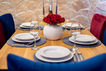 Elegant dining table set with white porcelain plates, wine glasses, and silverware, featuring a red rose centerpiece in a stylish white vase