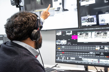 A television director in a broadcast control room, actively guiding a live production. Surrounded by multiple screens, the director wears a headset and gestures towards a video switcher.
