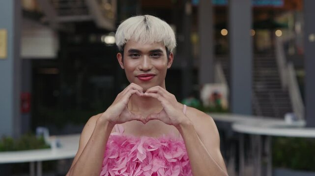 Stylish Genderqueer Person in Pink Top Posed Outdoors, Sending Love with a Hand Gesture. Cute Young Transgender Man Outside looking and Posing to the Camera. LGBT People Concept