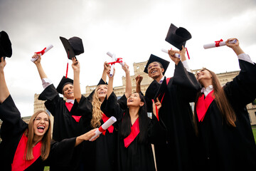 Joyful young graduates celebrate outside, wearing caps and gowns while raising diplomas in the air...