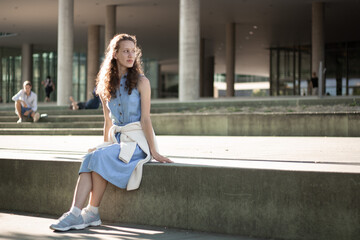 Girl sitting on steps in denim dress and sneakers. Student, studies, prospects, youth. Sunny weather. Admission.