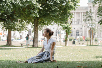 The girl sits thoughtfully on the grass in the park, dressed femininely and elegantly, looks sophisticated and looks away.