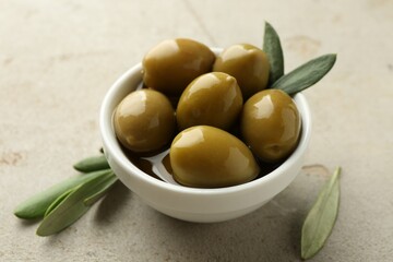 Delicious marinated olives in bowl and green leaves on grey table, closeup
