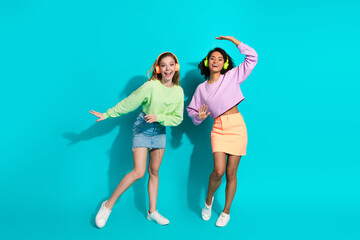 Two cheerful young women in stylish outfits posing with playful gestures on a teal background