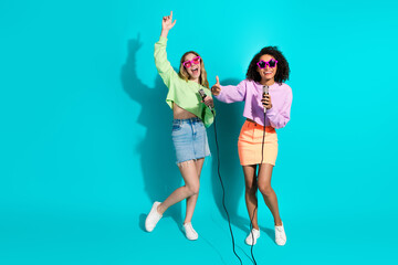 Two joyful young women singing karaoke and having fun in colorful casual outfits against a bright...