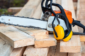 There is a chainsaw on a pile of firewood. The chainsaw is yellow and black, it is wearing yellow protective headphones