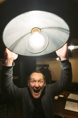 A joyful and cheerful man lifts a brightly glowing lamp with enthusiasm, showcasing his excitement in a cozy setting