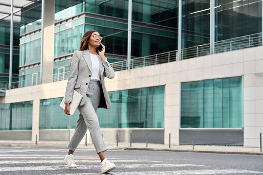Full body portrait of successful African American busy business woman having phone call outdoors. Middle age businesswoman in suit talking on smartphone, walking near office centre. Banner, copy space