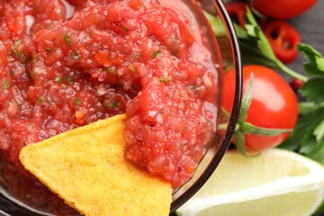 Spicy salsa sauce in bowl, nacho chip and ingredients on table, top view