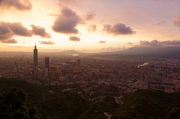 Aerial view of Downtown Taipei at sunset, with prominent 101 Tower standing among skyscrapers in Xinyi District and Tamsui River meandering by distant mountains under golden twilight sky in background