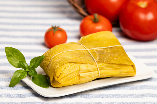 humita envuelta en hoja de choclo sobre un plato rectangular blanco decorado con una hoja de albahaca, sobre un mantel con lineas, al fondo tomates maduros que caen de un canasto