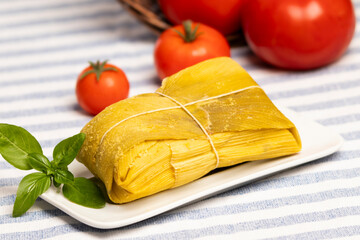 humita envuelta en hojas de maiz sobre un plato rectangular blanco decorado con una hoja de albahaca, sobre un mantel con lineas, al fondo tomates maduros de diferentes tamaños