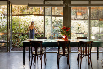 A woman in a sleek outfit stands on a contemporary balcony surrounded by lush greenery, enjoying the morning air. The open glass doors create a seamless connection between indoor comfort and outdoor