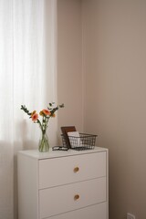 Colorful bouquet of beautiful gerbera flowers in glass vase, glasses, basket with books on white table indoors near the window.
