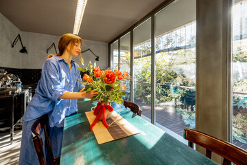 A woman in a relaxed blue shirt decorates a rustic dining table with a floral arrangement in a stylish kitchen. Natural light from large glass windows enhances the warm and inviting atmosphere