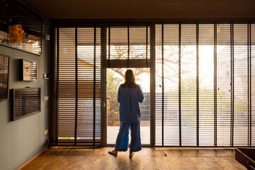 A woman in a relaxed blue outfit stands by a large window with wooden blinds, gazing outside. The warm morning light fills the stylish interior, creating a serene, contemplative, and cozy atmosphere