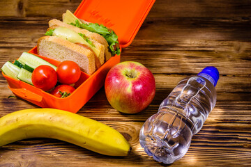 Ripe apple, banana, bottle of water and lunch box with sandwiches, cucumbers and tomatoes on wooden...