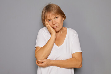 A woman is standing against a gray backdrop, resting her hand on her cheek, showing signs of deep...