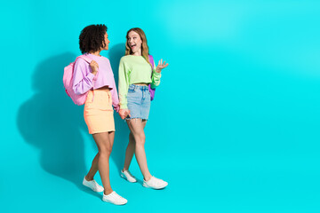Obraz premium Two young women walking and chatting in trendy casual outfits against a vibrant turquoise background