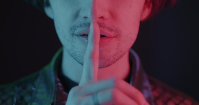 Close-up of man touching mouth with finger shushing asking for silence in neon light on dark background. People and hand gesture concept.