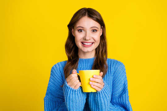 Smiling young woman holding a yellow mug against a vibrant yellow background in a casual blue sweater