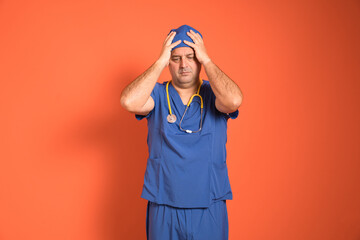 Overworked surgeon suffering from headache or stress, wearing blue scrubs and stethoscope, touching his head with hands on orange background