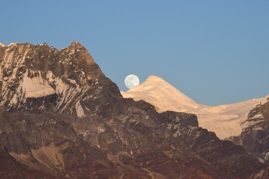 "A breathtaking view of the full moon rising behind a majestic snow-capped mountain. The golden light of the setting sun highlights the rugged mountain peaks, creating a stunning contrast 