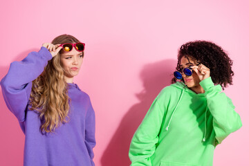 Two stylish young women in casual sweatshirts posing playfully with sunglasses against a vibrant pink background