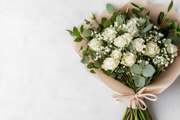 Obraz premium A delicate bouquet features elegant white roses, accented with sprigs of lush greenery and tiny white flowers, wrapped in soft beige paper and tied with a ribbon. The composition exudes freshness.