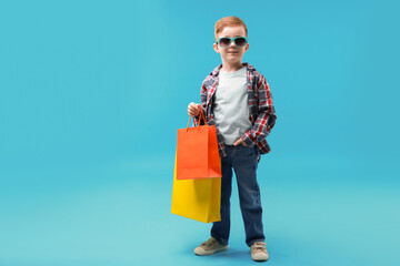 Cute little boy with shopping bags on light blue background. Space for text