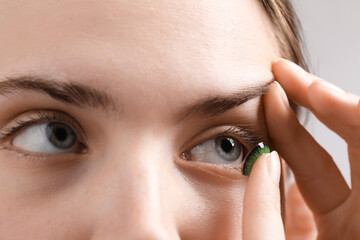 Woman putting in green color contact lens on grey background, closeup