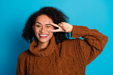 Smiling young woman in a brown knitted sweater making a fun gesture with a blue background